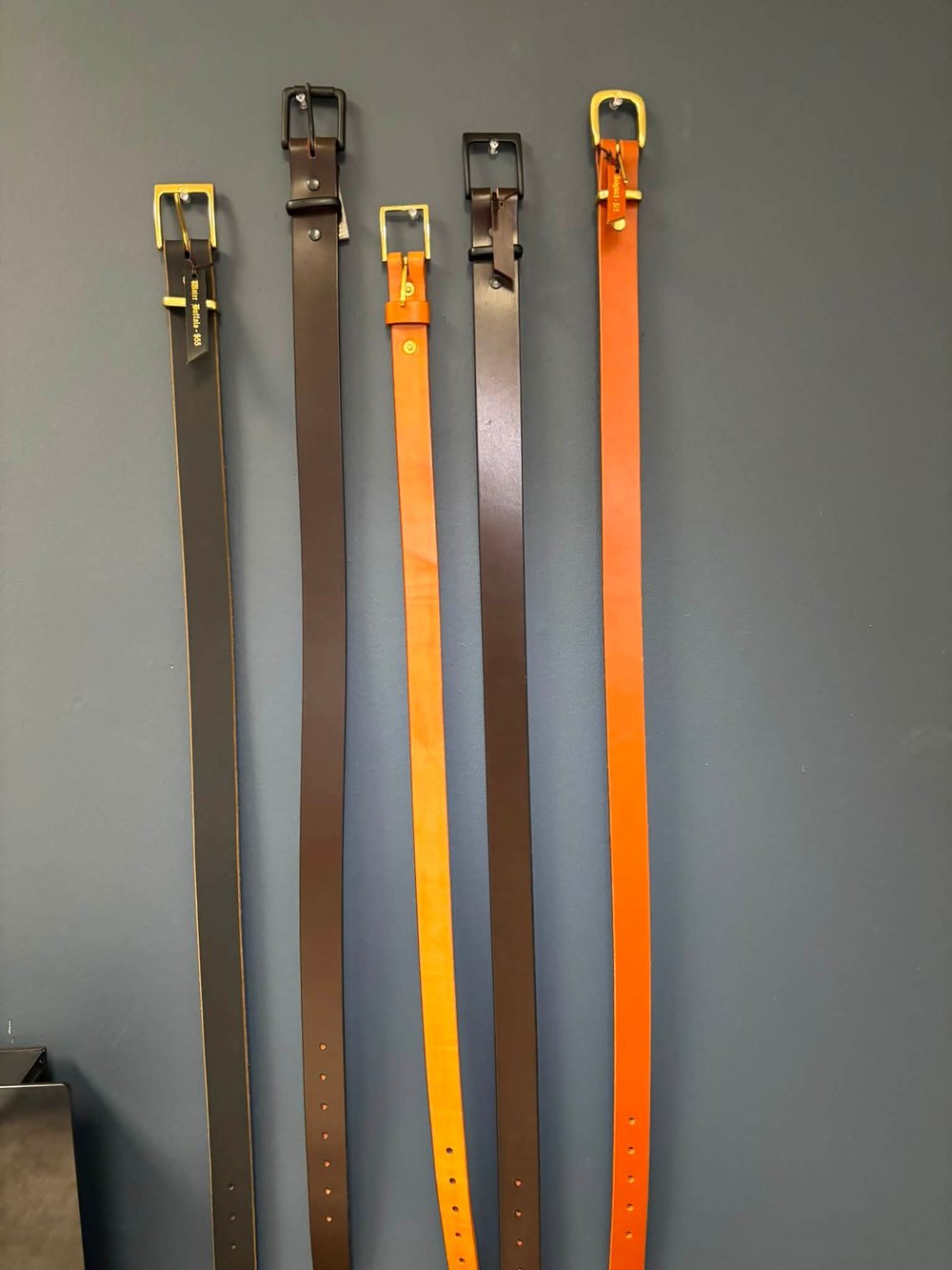 Four finished belts hanging in a row, brass buckles, in gray, tan, dark brown and orange