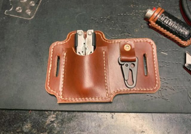 A belt sheath carrying a multi-tool and a folding knife side by side, stitched and burnished, resting on the cutting mat