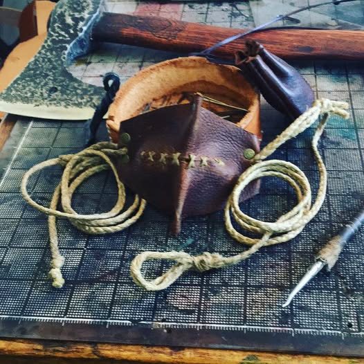 A hand-stitched leather sling with its braided cords coiled beside a hand axe